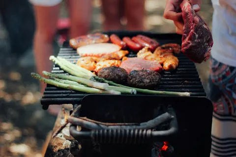 Mehrere Personen stehen am Holzkohlegrill mit Würstchen, Burgern, Hähnchen und Spargel, eine Hand legt ein Steak auf